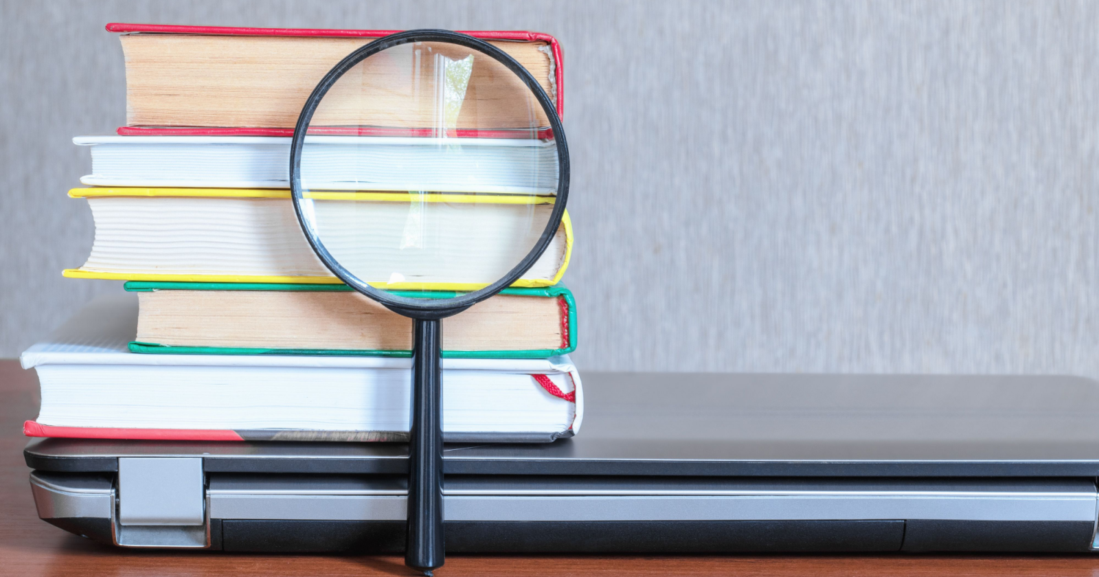 A magnifying glass in front of a stack of books and a closed laptop on a wooden desk, primed for search results analysis.
